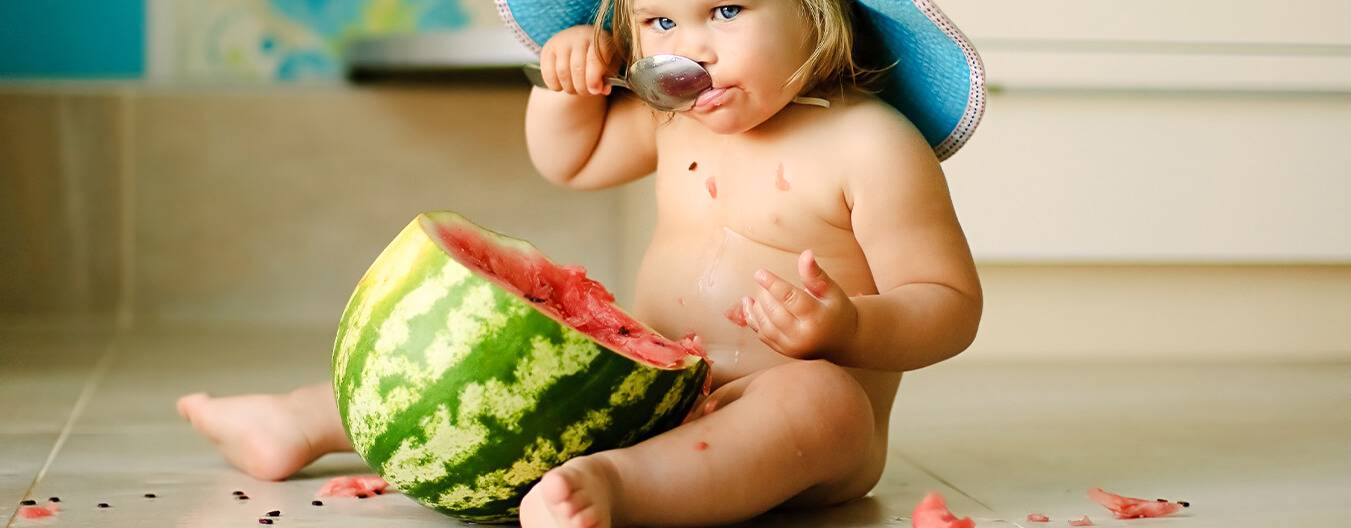 Baby with diapers sitting over tile floor eating a watermelon with spoon